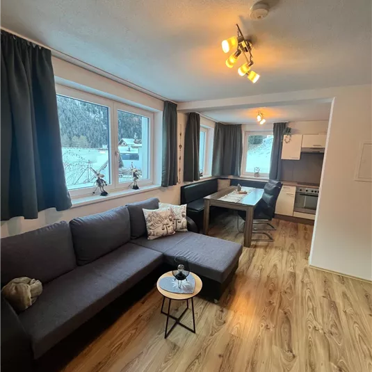 A modern living room with a gray sofa and a small table. Large windows provide plenty of daylight and a view of the surroundings.