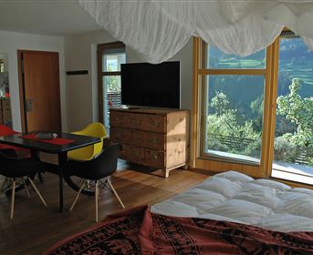 Ein lichtdurchflutetes Zimmer mit großen Fenstern und Blick auf die Natur. Es ist modern eingerichtet mit einem Tisch, bunten Stühlen und einem gemütlichen Bett.
