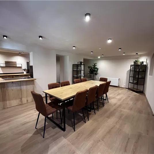 A modern meeting room with a long wooden table and chairs. Behind the table is an open kitchen and some plants in the corner.