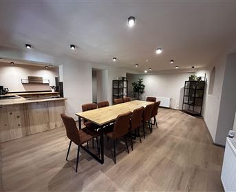 Ein moderner Tagungsraum mit einem langen Holztisch und Stühlen. Hinter dem Tisch befindet sich eine offene Küche und einige Pflanzen in der Ecke.