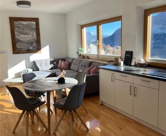 A modern living and dining room with large windows and a view of the mountains. The space is brightly lit and cozily furnished.