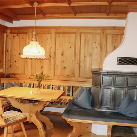 A cozy dining area made of wood with a bench and a table. A decorative lamp and a traditional stove contribute to the warm atmosphere.