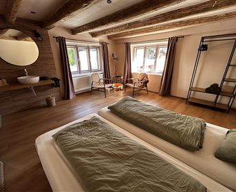 Ein modernes Schlafzimmer mit zwei Betten und einem warmen Holzfußboden. Große Fenster lassen viel Licht herein und schaffen eine einladende Atmosphäre.