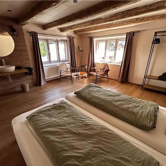 Ein modernes Schlafzimmer mit zwei Betten und einem warmen Holzfußboden. Große Fenster lassen viel Licht herein und schaffen eine einladende Atmosphäre.