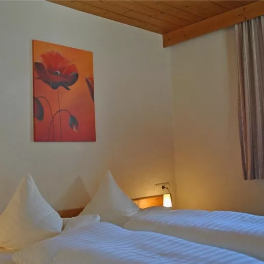 A cozy room with two beds and light wood decor. A picture of red flowers hangs on the wall, and the light is warm and inviting.