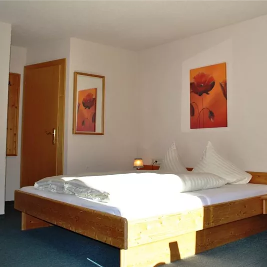 A cozy room with a double bed and wooden-colored furniture. Pictures of flowers hang on the walls, and there is pleasant lighting.