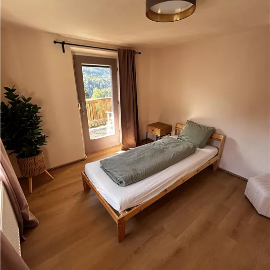 Ein einfaches Schlafzimmer mit einem Einzelbett und einem kleinen Nachttisch. Durch das Fenster sieht man die Natur draußen.