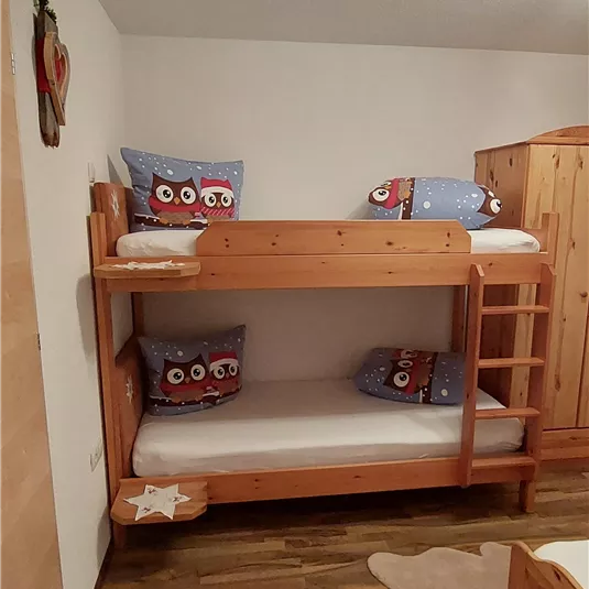Ein gemütliches Kinderzimmer mit einem Etagenbett aus Holz. Die Betten sind mit Kissen in Eulenmotiven dekoriert und der Boden ist mit einem Teppich geschmückt.