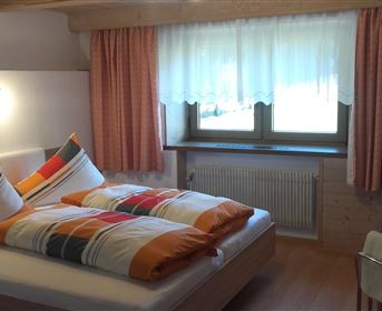 Ein gemütliches Schlafzimmer mit einem Doppelbett und bunten Kissen. Große Fenster lassen viel Tageslicht herein.