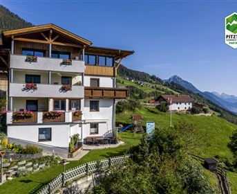 Ein gemütliches Ferienhaus in den Bergen mit blühenden Balkonen. Im Hintergrund erstrecken sich grüntönige Hügel und hohe Berge.